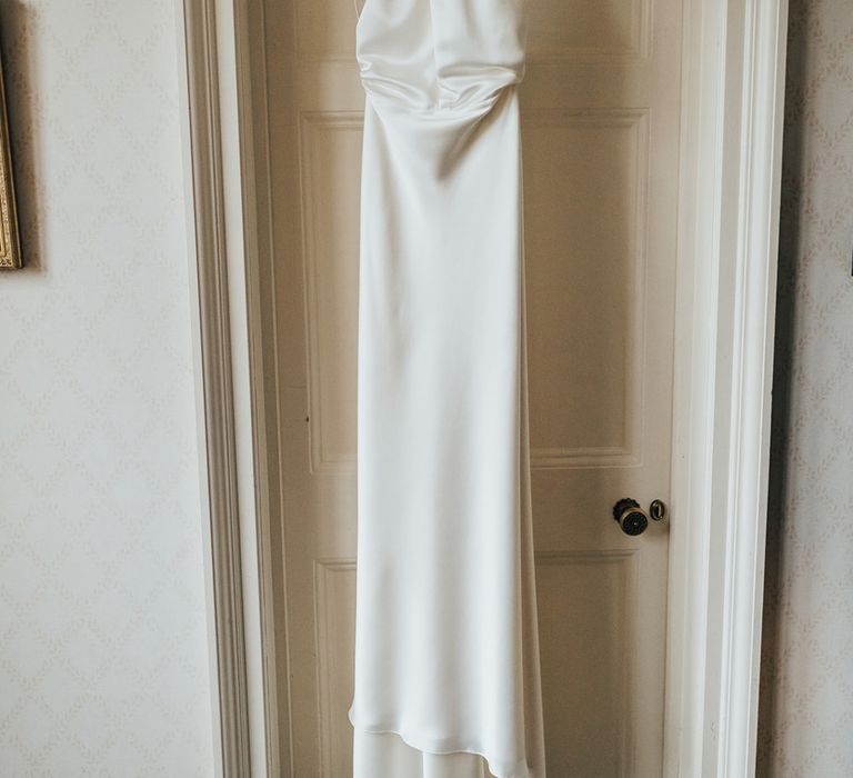 Halterneck satin Halfpenny London wedding dress hanging from doorway before church wedding ceremony