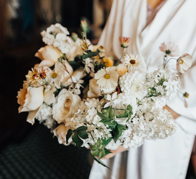 Bride in white satin dressing gown holding mixed white and green bridal bouquet with roses for wedding in Hertfordshire