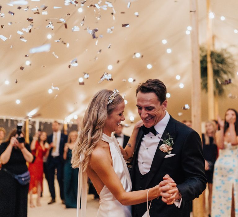 Bride in satin halterneck wedding dress and bridal headband has first dance with groom in black tie as silver confetti falls at sperry tent wedding reception