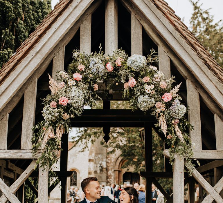 Bride and Groom leaning on the gate at the church with pretty blue hydrangea and pink rose flower arch 