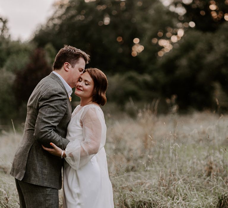 Golden house portrait with groom in a grey check suit kissing his bride in a sheer long sleeve wedding dress 