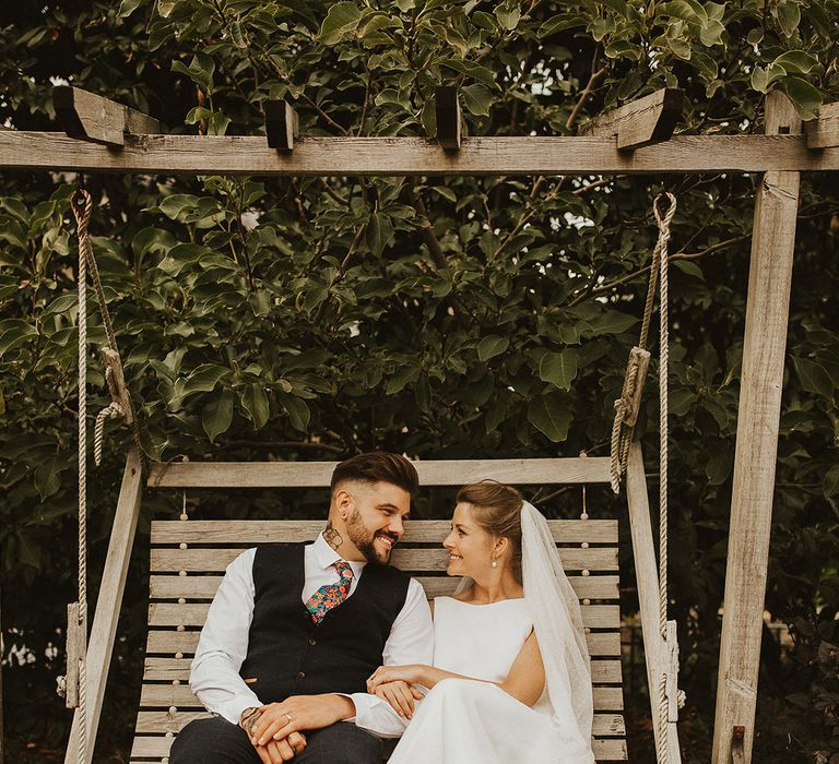 Bride and groom portrait sitting on a wooden swing with groom in a waistcoat and floral tie and bride in bow front sandals and cathedral length veil 