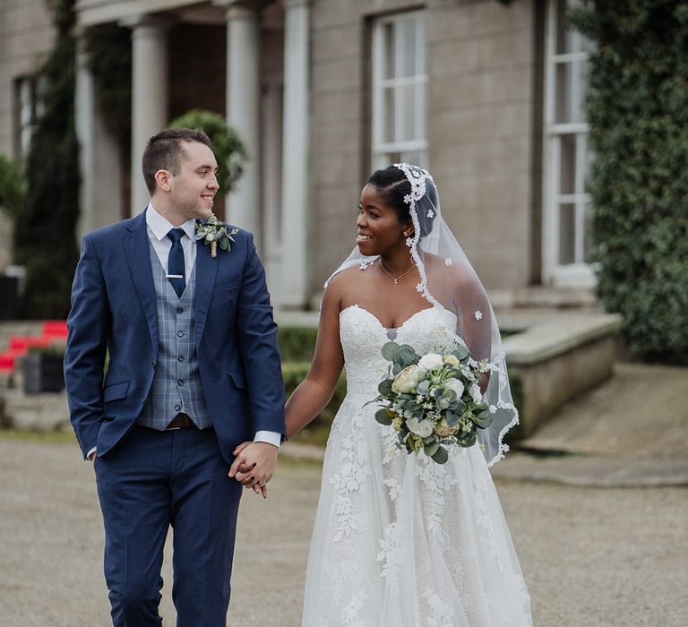 Bride in sweetheart neckline wedding dress with lace edge veil smiles with Groom in Navy three piece suit at their wedding ceremony  