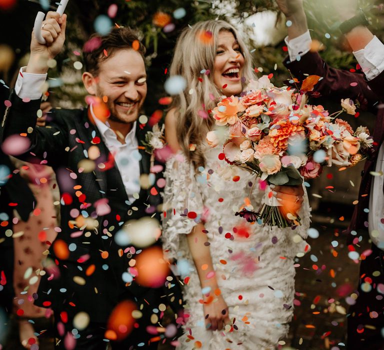 Bride in floral applique wedding dress holding peachy bridal bouquet smiles whilst walking with groom in dark suit as guests throw multicoloured confetti 