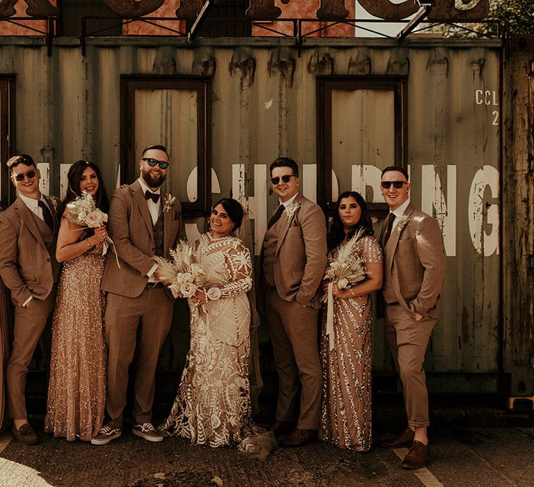 wedding party photograph standing n front of industrial containers with groomsmen in tweed suits, bridesmaids in blush dresses and bride in a lace wedding dress 