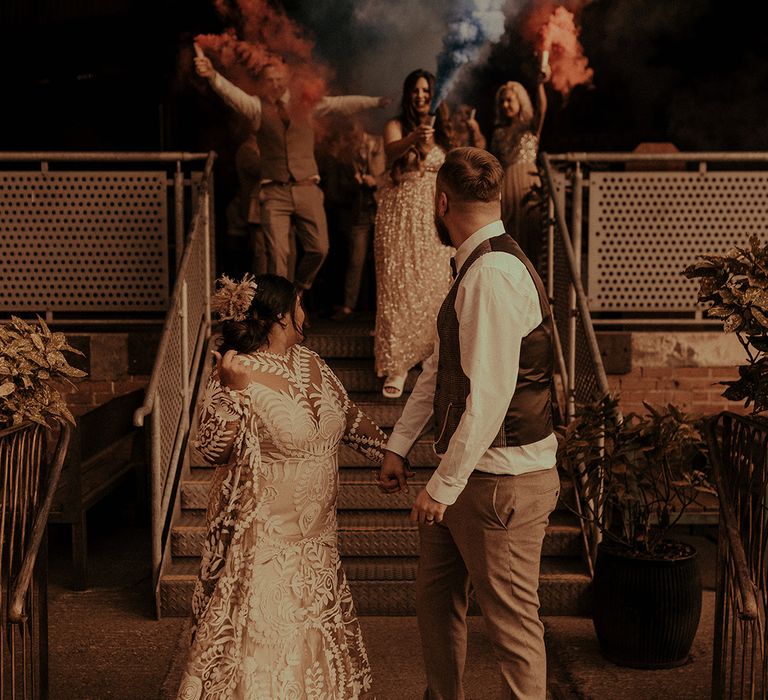 Bride in a boho lace wedding dress holding hands with her groom in a tweed waistcoat and vans with their wedding party behind them holding smoke flares 