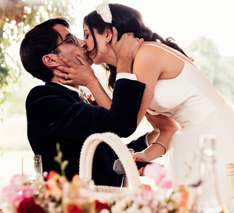 Bride in thin strap wedding dress with tulle skirt and feathered headband kisses groom in dark suit after talking speech during wedding breakfast