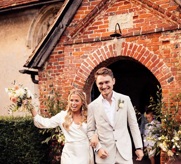 Bride in white satin dress with wrap top holding pink and yellow wedding bouquet walks out of church arm in arm with groom in light grey suit, open white shirt and brown suede shoes