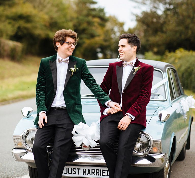 Grooms hold hands seated together on vintage baby blue wedding car transportation with white ribbons and bow ties