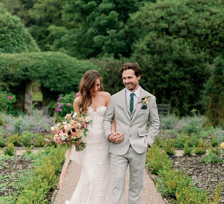 Bride in strapless lace wedding dress and holds hand with groom in bespoke light grey suit and green tie