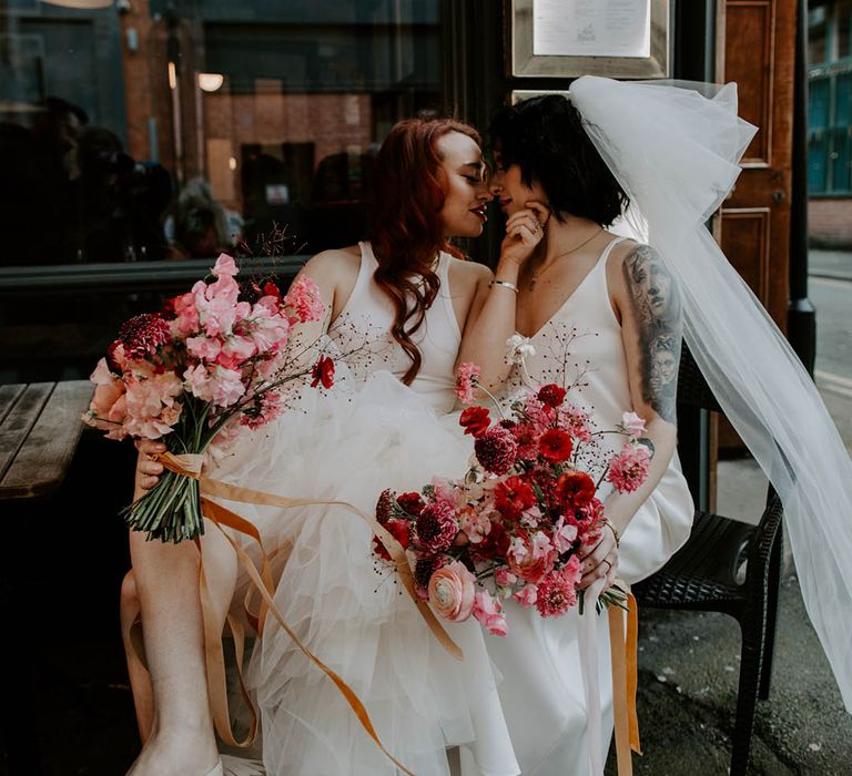 Two brides sitting and kissing in a satin slip wedding dress with big bow tulle veil and high low layered tulle wedding dress