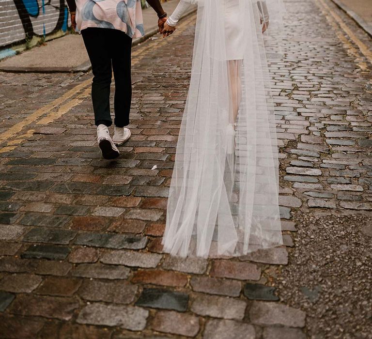 Groom in a patterned shirt and Converse trainers holding hands with his bride in a short wedding dress with long veil