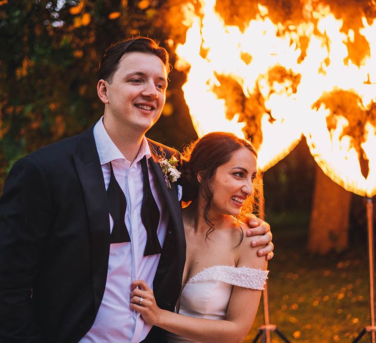 Bride in sparkly Pronovias wedding dress with groom in black tie stand in front of flame hearts