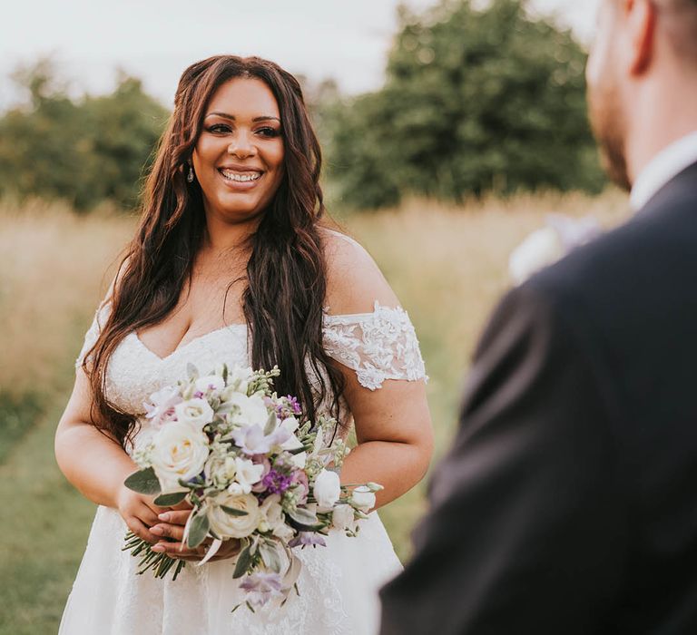 Bride with long brown hair in off the shoulder lace wedding dress holds a purple and white bouquet as she smiles at the groom