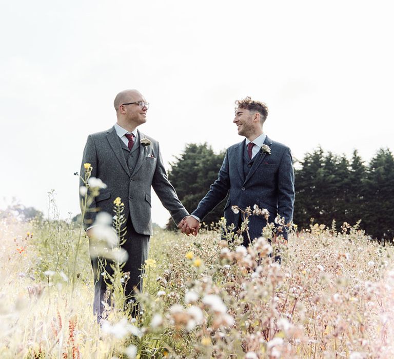 Groom in blue suit and red tie holds hands with groom in grey suit with red tie in a field together 