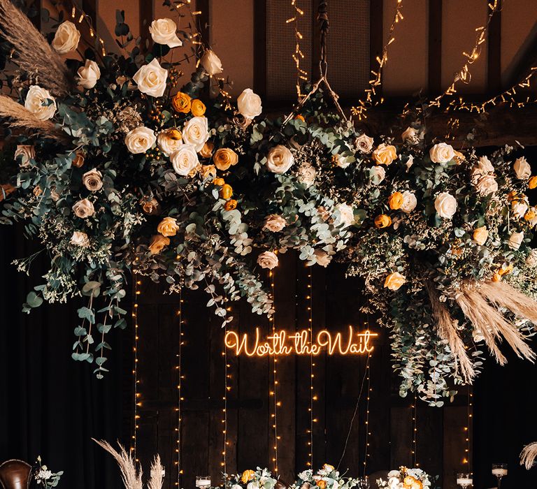 Wooden banquet top table with orange and white flower cloud, dried flowers, candle lanterns in rustic styling with neon 'Worth the Wait' sign 