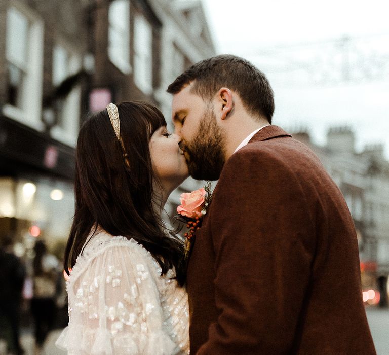 Bride in Needle and Thread sparkly top and gold headband kisses the groom in brown suit 