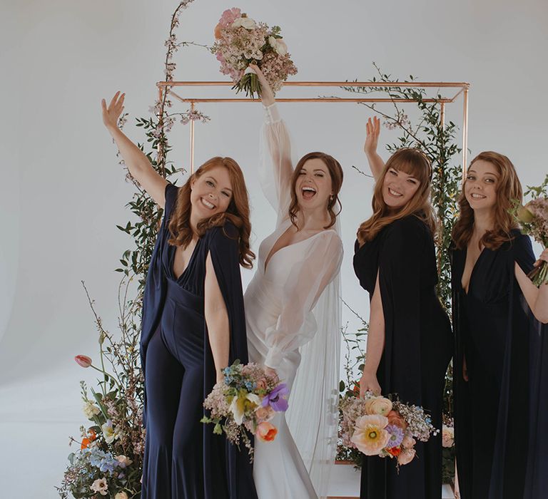 Bride in Suzanne Neville dress with custom sheer sleeves posing with bridesmaids in navy jumpsuits and cape arm detail 