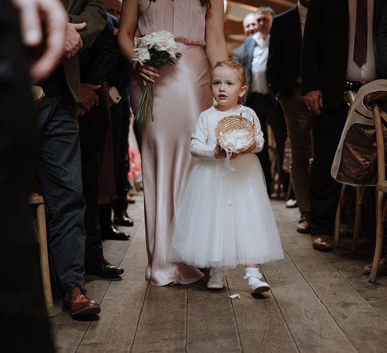 Flower girl in white pearl cardigan and tulle dress with frilly socks pours out petals from wicker basket 