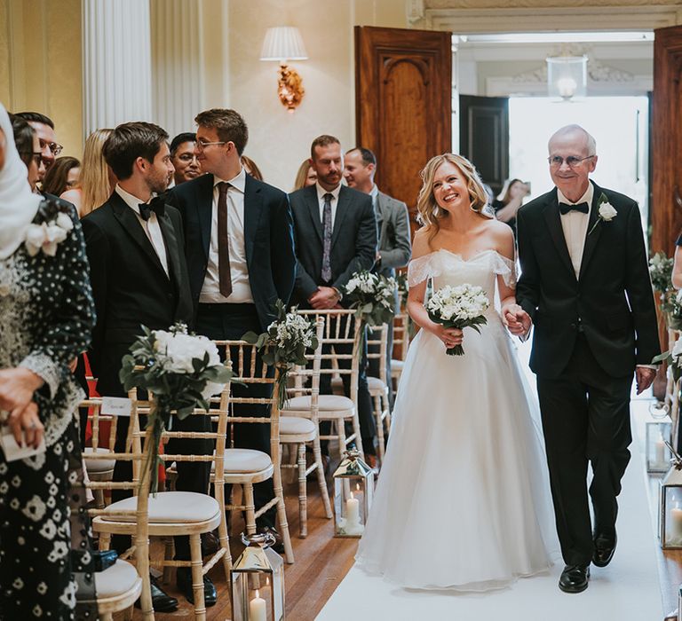 Father of the bride in black tie walks the bride in off the shoulder wedding dress down the aisle 