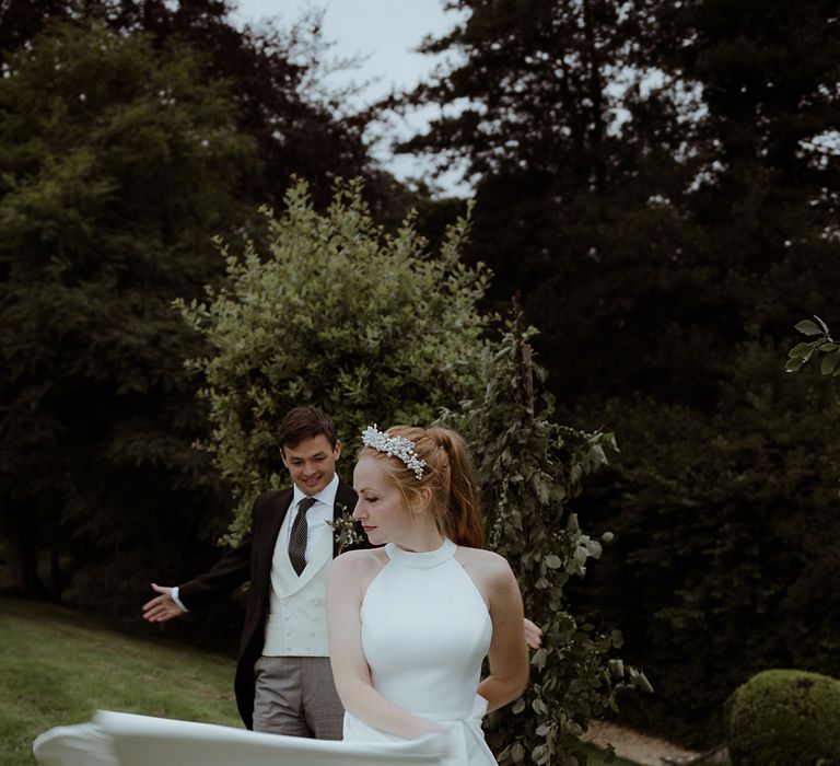 Bride spins around in a halter neck pleated skirt wedding dress and pearl crown with groom watching in the background 