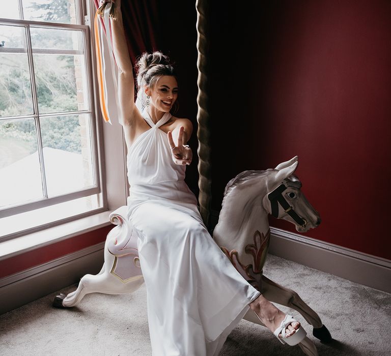 Bride in a satin halter neck wedding dress and platform shoes sitting on a unicorn merry-go-round horse at Garthmyl Hall 