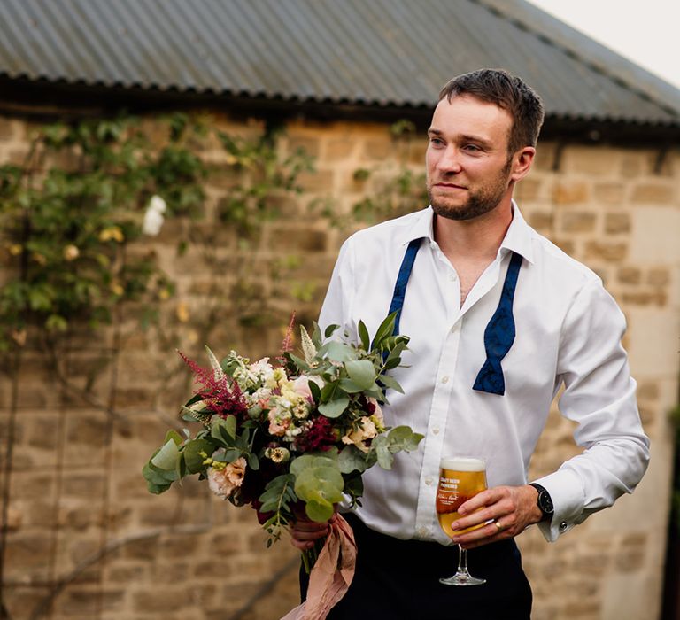 Groom in white shirt and undone blue bow tie holding glass of beer and the red and white bridal bouquet