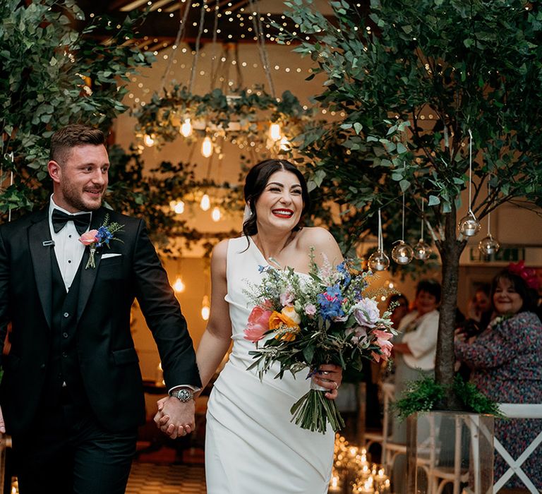 Groom in black tie and bride in one shoulder wedding dress with red lipstick walk out of their ceremony holding hands with the bride holding a colourful bouquet that matches the groom's buttonhole