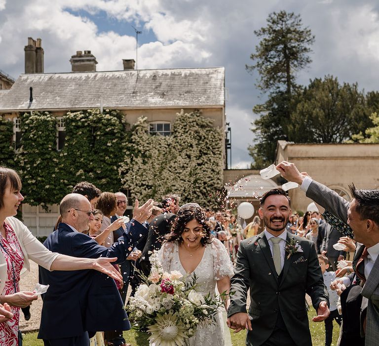 Bride & groom walk through confetti outdoors whilst holding meadow floral bouquet for Greek blessing wedding ceremony