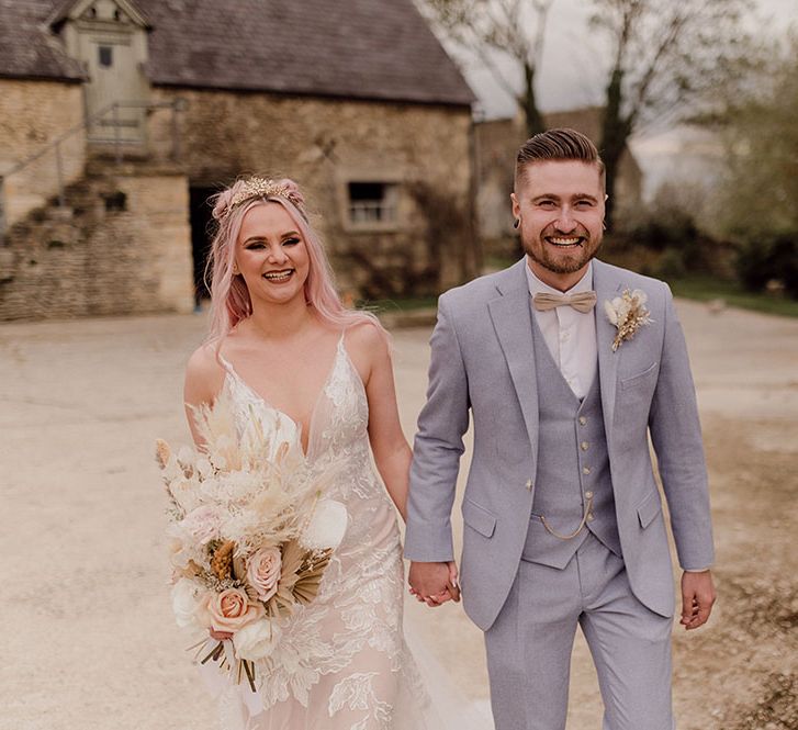 Bride wearing Jane Hill fitted wedding dress and holding dried floral bouquet walks with her groom in pale grey three piece suit and nude bow tie