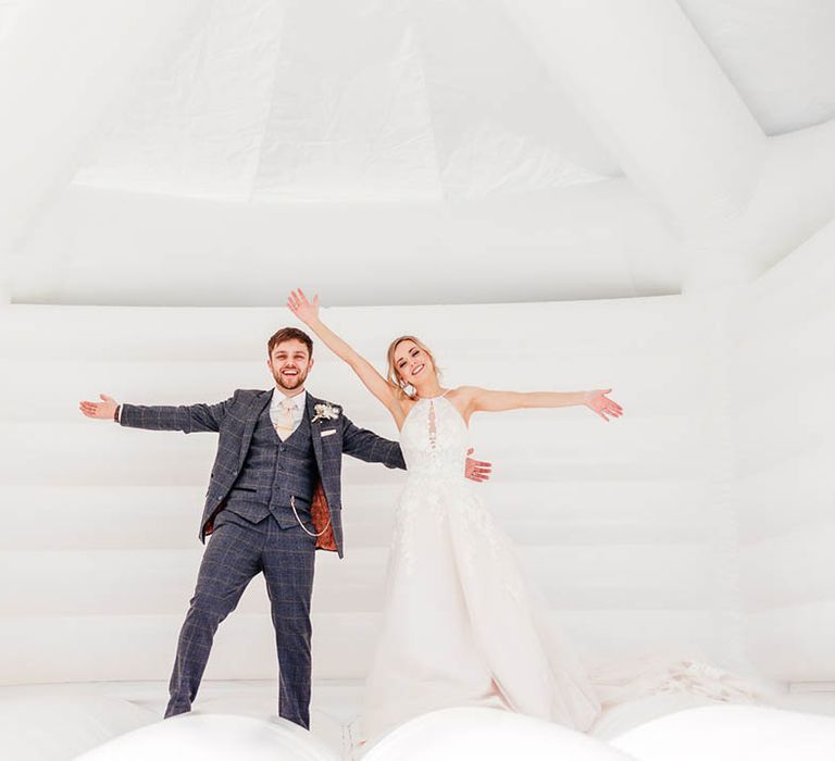 Bride and groom smile and spread out their arms together on their white wedding bouncy castle 