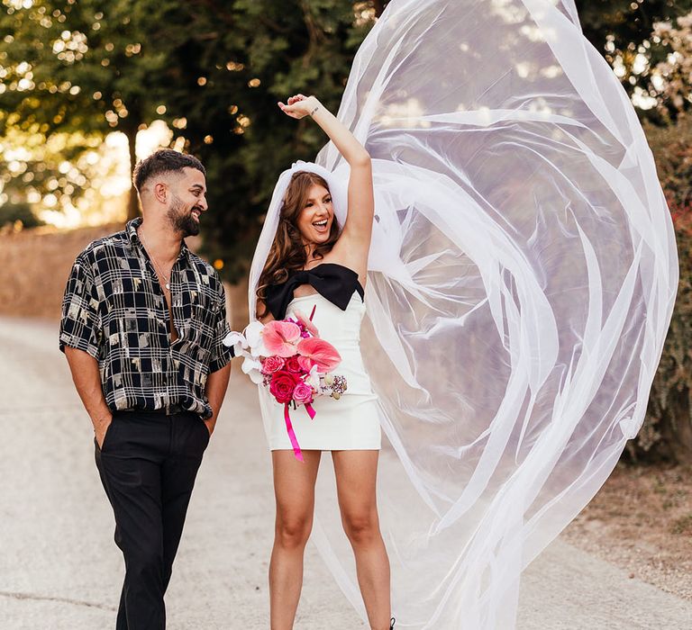 Bride wears mini dress with black bow and holds brightly coloured floral bouquet as she throws out her veil beside her 