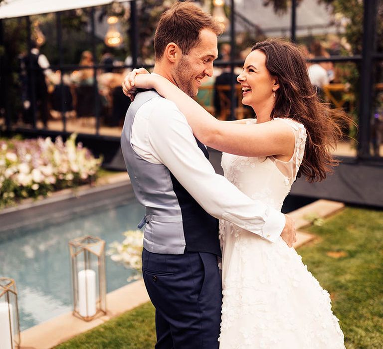 Bride and groom embrace in front of mini pool in next to their glass marquee reception 