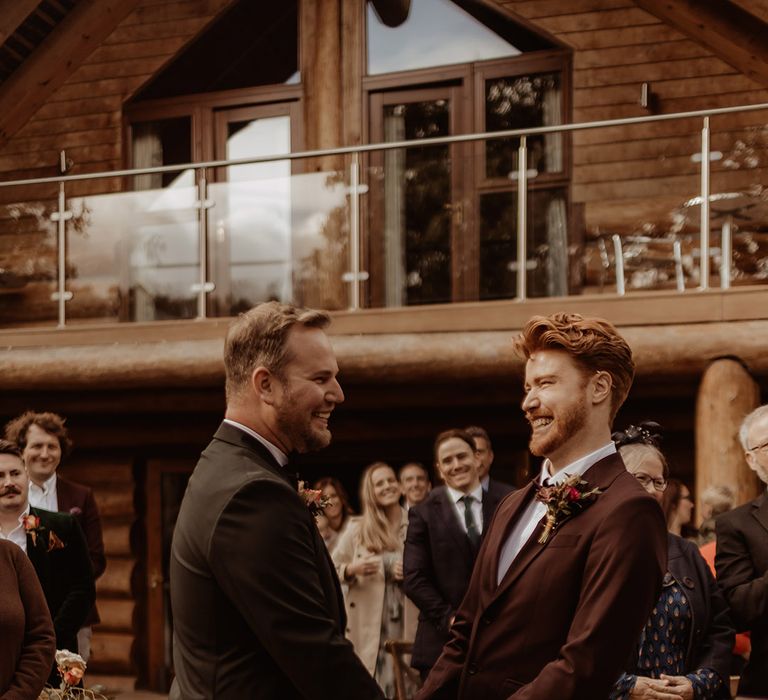 Groom in black tie and groom in a burgundy suit stand holding hands at the altar for their outdoor rustic ceremony