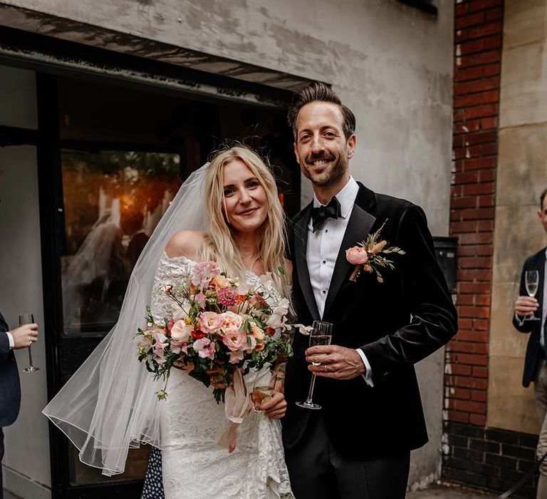 Bride in lace boho two piece, colourful mixed floral bouquet, gold strappy heels and short veil and groom in black tux with bow tie and boutonniere