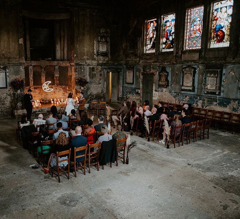 The wedding ceremony taking place at industrial venue in London featuring a moon neon sign at the centre of the aisle