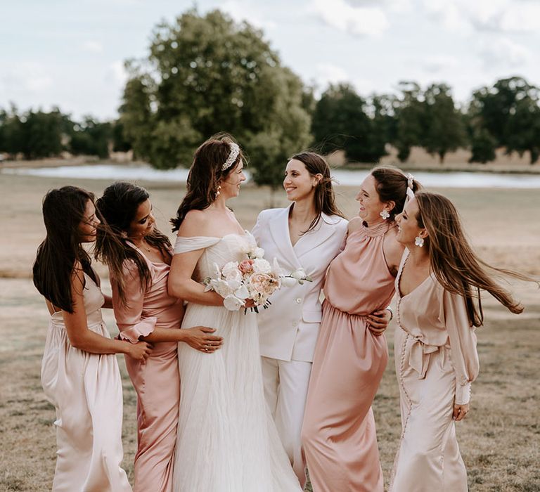 The bride in a white bridal suit smiling with the bride in an off the shoulder wedding dress with bridesmaids in pink and champagne coloured dresses 