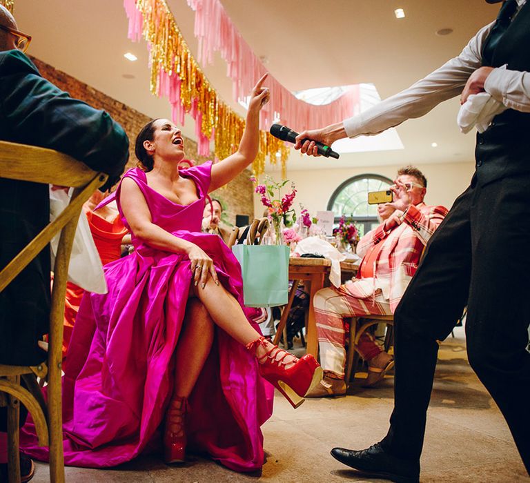 Bride in off the shoulder front ruching pink wedding dress and Vivienne Westwood elevated red patent leather platform heels with shoe-string fastening singing into microphone at wedding entertainment part of the reception