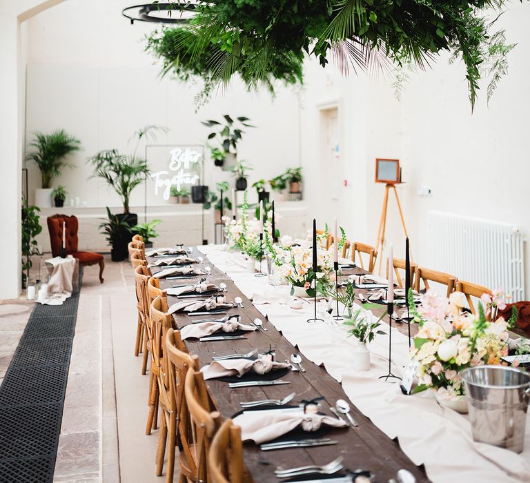 Reception room of The Orangery wedding venue with neutral off-white table runners, rose floral arrangements, pink and black tapered candles, and suspended foliage decorations