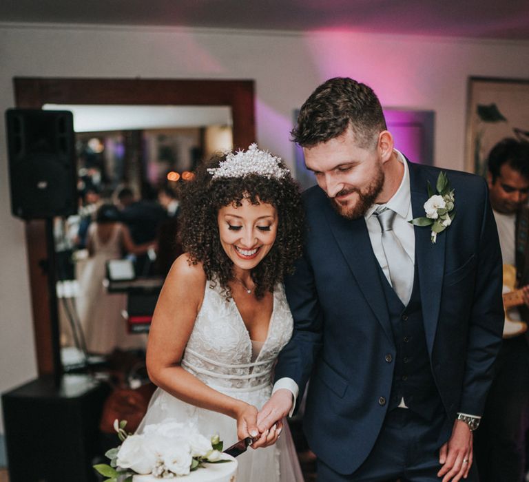 Bride in pearl embellished bridal crown cuts wedding cake alongside her groom in three piece suit 