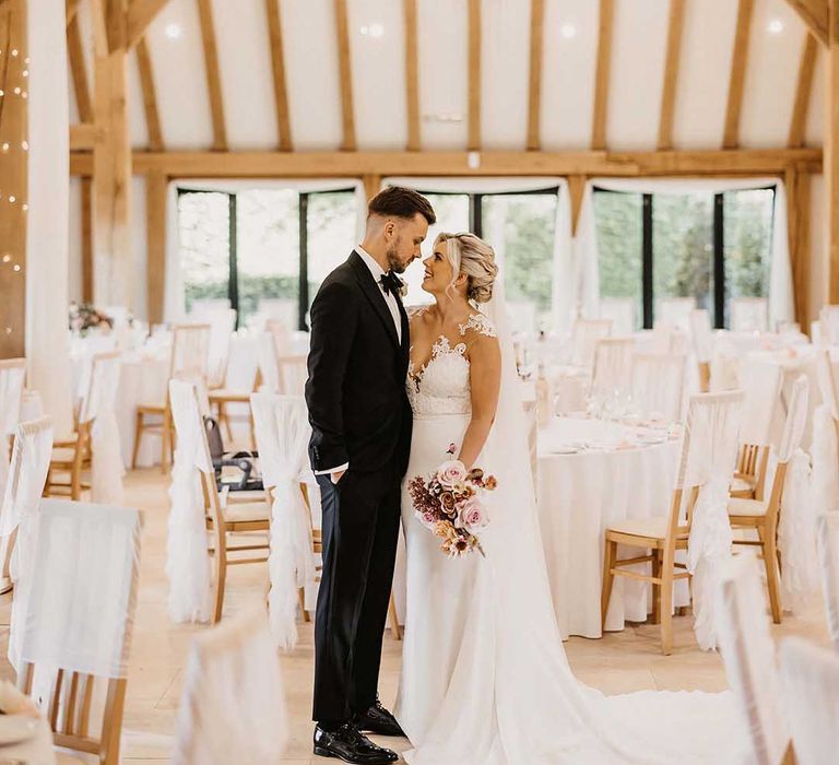 Bride in lace wedding dress with sheer, lace top and clusters of lace flowers on the shoulders and plunge neckline in the reception room of Old Kent Barn with chandeliers, fairy lights and wedding drapes 