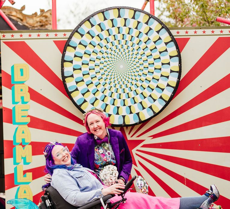 Bride in purple velvet blazer and black suit trousers with purple bowtie, pink patterned pocket square and colourful patterned shirt and bride in wheelchair wearing personalised denim jacket, pink wedding dress and blue tights posing in front of kaleidoscope sign at theme park wedding 