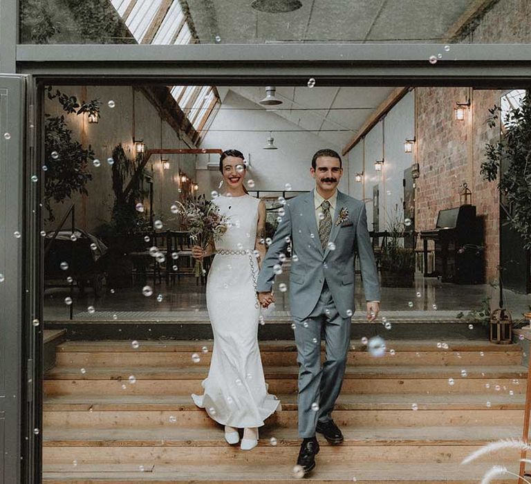 Lisa in pre-loved fitted wedding dress walks alongside Gabriel in powder blue Gucci suit through bubbles 