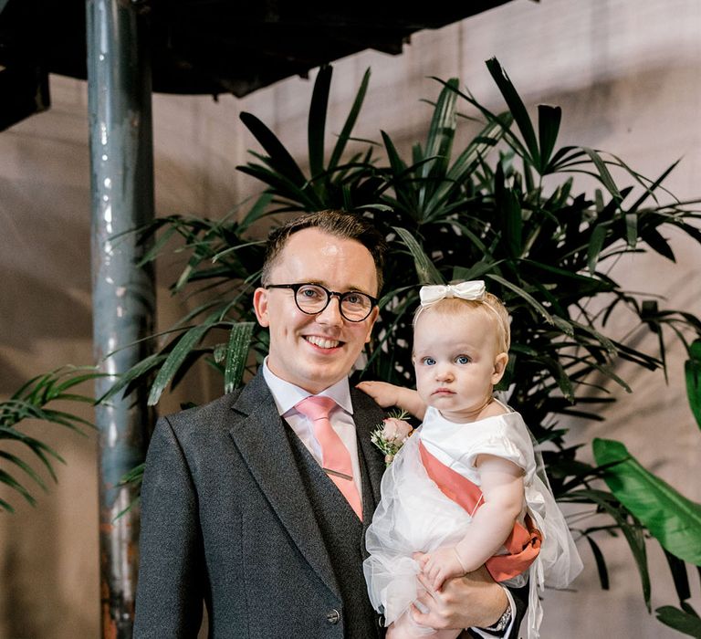 Groom wears grey suit jacket and pale pink tie as he holds his daughter in white dress with pink sash 