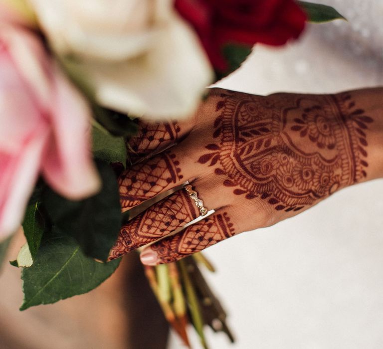 intricate bridal henna across bride's hand holding red, white and pink rose bouquet 