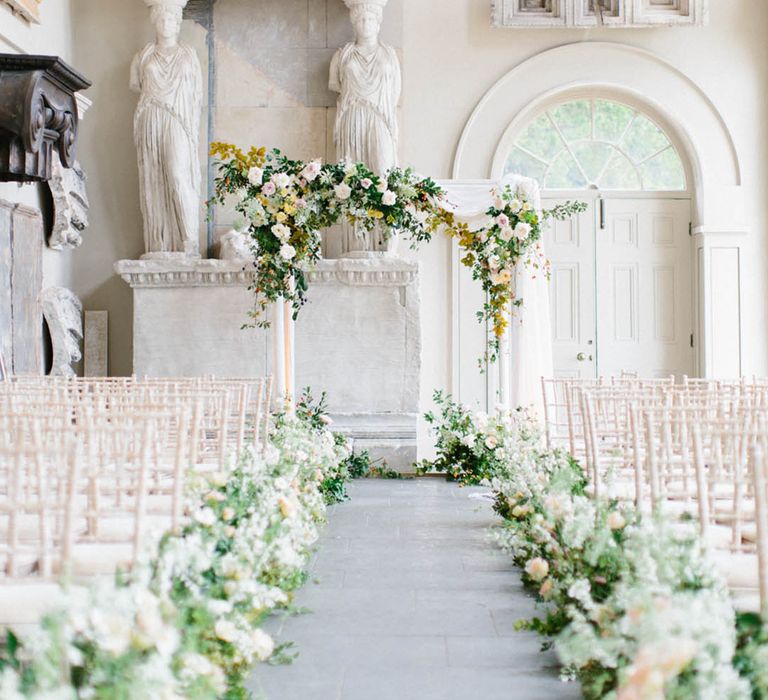 White wedding flowers lining the aisle with metal arch altar decoration with white drapery for indoor aisle decor 