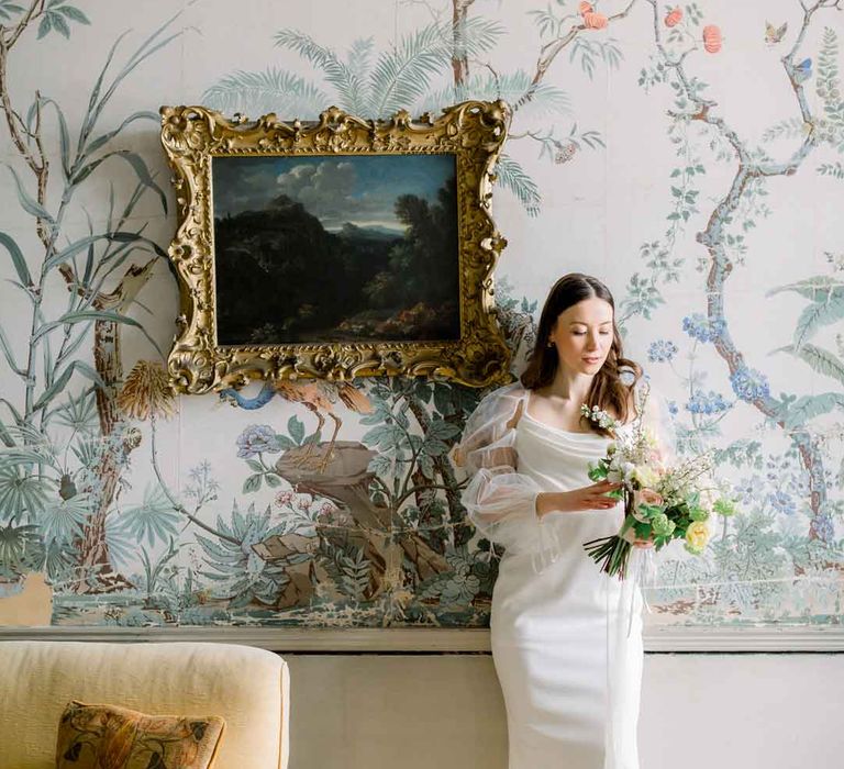 Bride holding classic bridal bouquet wearing long sheer puff sleeve satin wedding dress with cowl neck standing by yellow velvet sofa at St Giles House