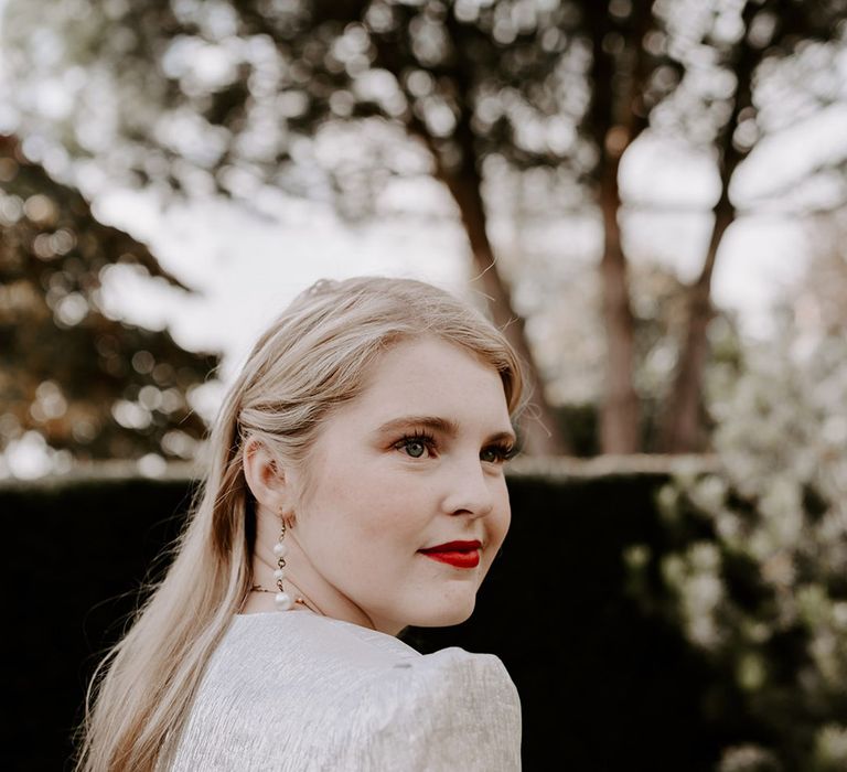 Bride in a The Vampire's Wife silver metallic wedding dress with a red rose bouquet, red lipstick, and pearl earrings 