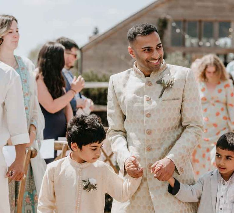 Groom in cream and gold Indian sherwani and white garden rose boutonniere walking down the aisle with two page boys at Gujarati wedding ceremony 