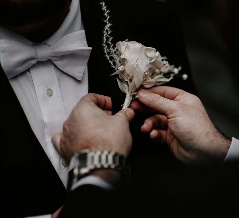 Groom wearing silver watch fastening groom's white garden rose, lavender twig and dried foliage boutonniere at LGBTQ+ London wedding